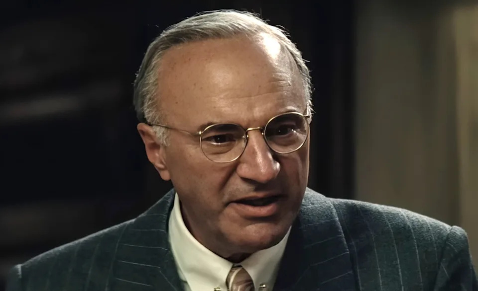 An older man in a pinstripe suit and glasses speaks intently in an indoor setting