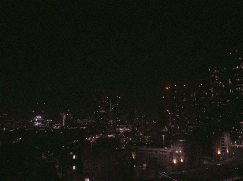 A dark cityscape at night with scattered lights from buildings and windows; tall skyscrapers are visible against the night sky.