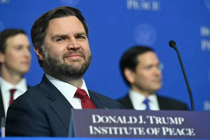 A man in a suit and red tie sits at the Donald J. Trump Institute of Peace event. Other attendees are blurred in the background