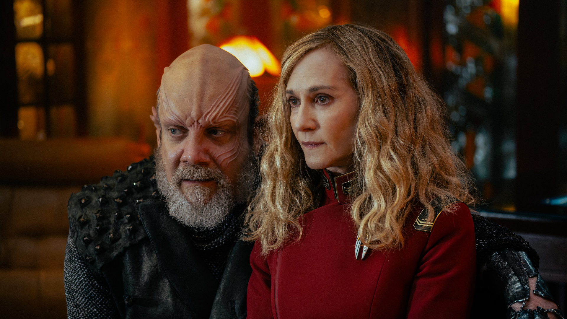 a humanoid alien with x's and o's shaved into the side of his head and a raised cranium kneels beside a woman with curly blonde hair in a red officer's uniform, both of them looking off into the distance inside a richly appointed office