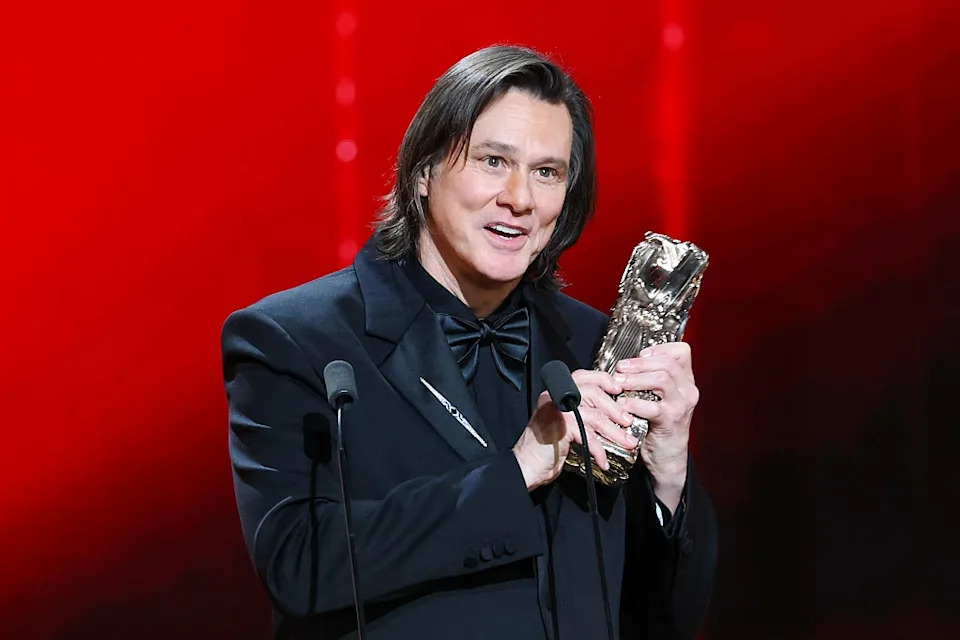 Person in a formal suit holding a silver bear-shaped award, speaking into microphones on stage
