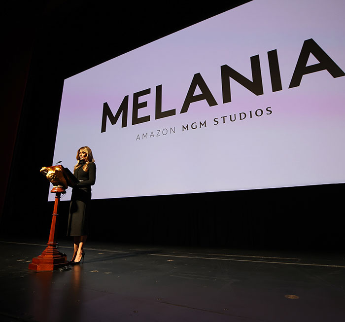 Speaker standing at podium on stage in front of large screen displaying Melania Amazon MGM Studios logo during theater event. Speaker standing at podium on stage in front of large screen displaying Melania Amazon MGM Studios logo during theater event.