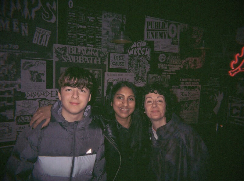 Three people, two women and one teenage boy, smile and pose together in a dimly lit room with walls covered in music and band posters, including ones for Black Sabbath and Acid Witch.