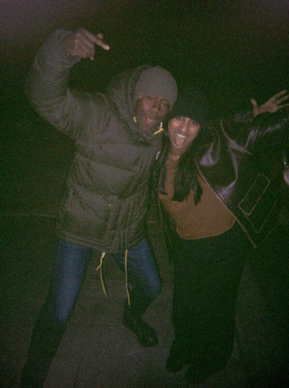 Two people in winter jackets and hats pose playfully outdoors at night, smiling and making expressive gestures toward the camera. The background is dark and not visible.