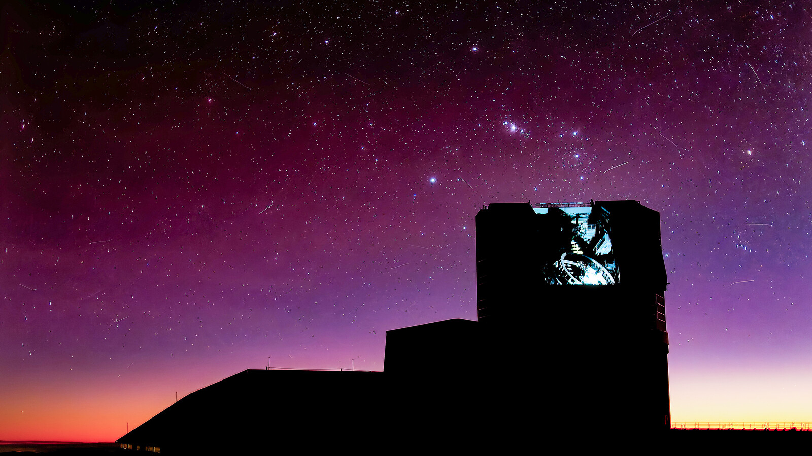 The Vera Rubin Observatory, a large building, is silhouetted against a bright purple night sky