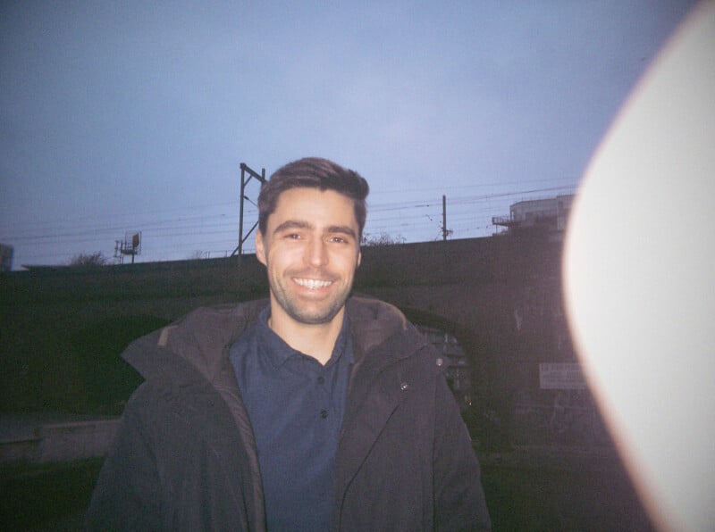 A man with short brown hair and a beard smiles at the camera, standing outdoors in front of a dark bridge at dusk. He wears a dark jacket and a navy shirt. A finger partially covers the right side of the photo.