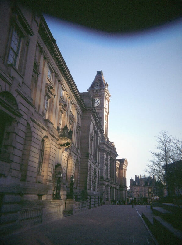 A historic stone building with a clock tower is shown at sunset. The sky is clear, a few people walk along the path, and bare trees line the street. The top of the photo is slightly darkened by a shadow.