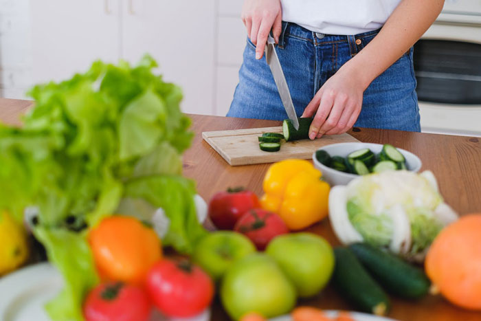 Woman slicing cucumber on a cutting board in kitchen surrounded by fresh vegetables, symbolizing parenting and family dynamics.