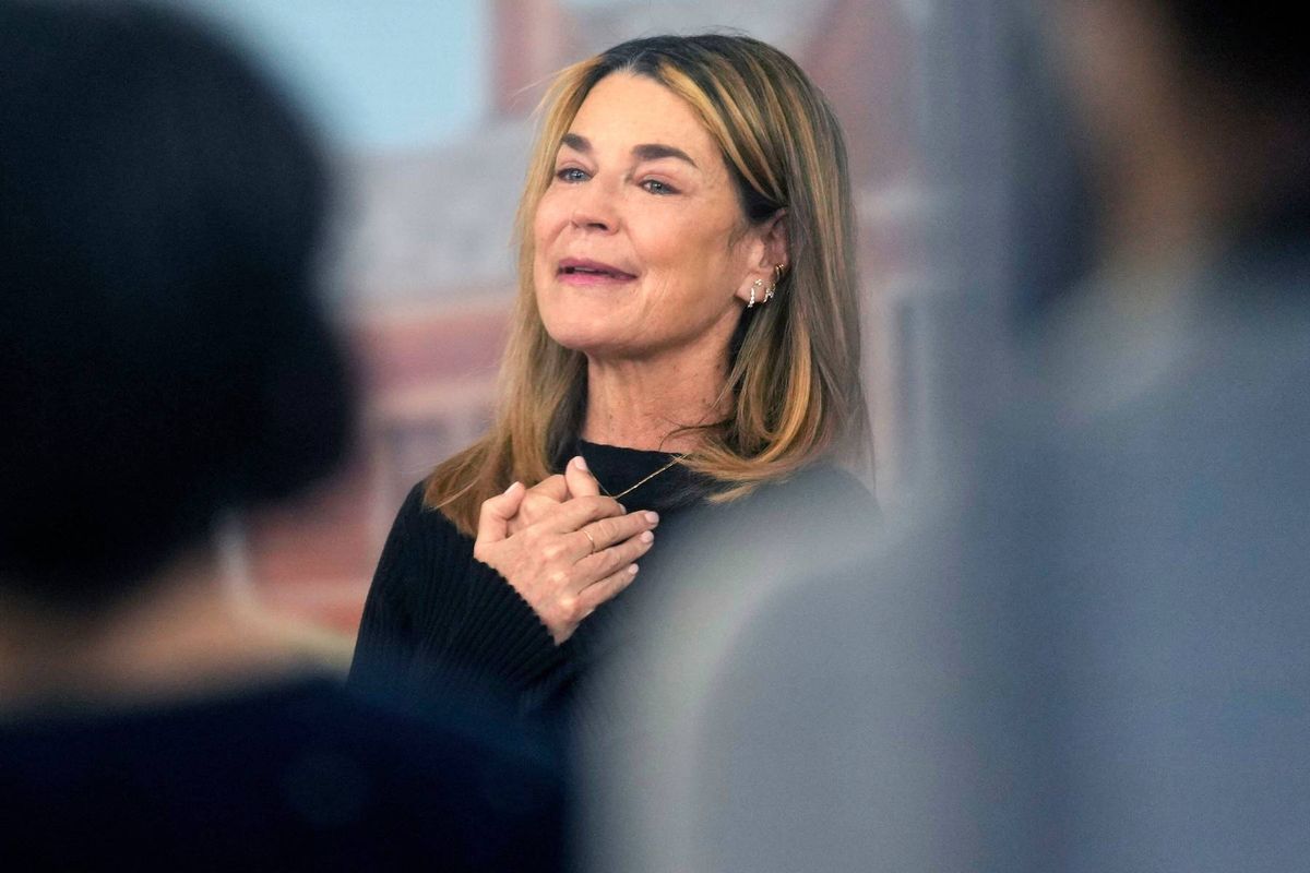 An individual with blonde hair, dressed in a black top, appears to be engaged in an animated discussion with others in a room. The person's facial expression suggests a moment of emphasis or emotion during the conversation.