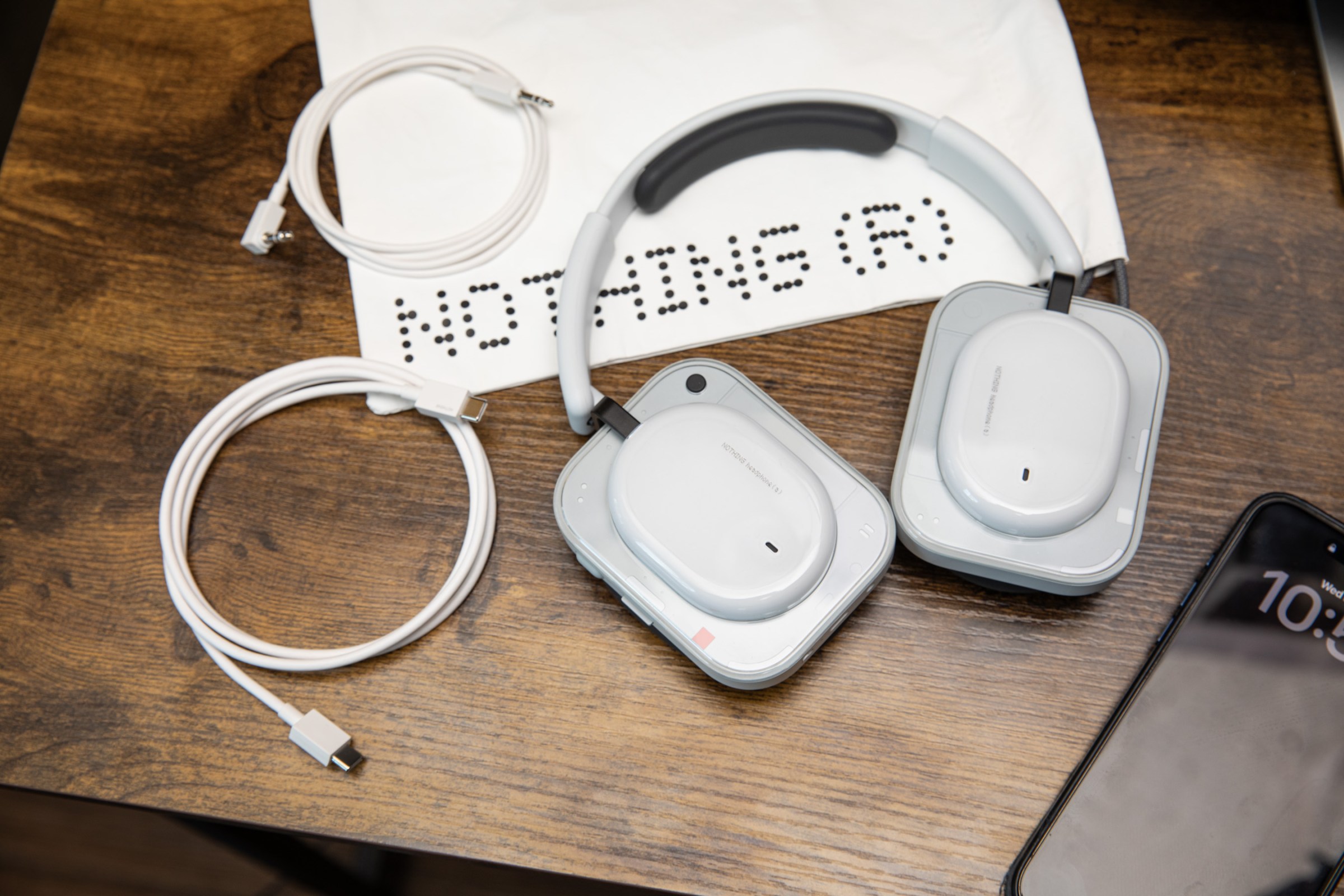 The Nothing Headphone A with its carrying bag, 3.5mm cable, and USB-C cable on a wooden desk with an iPhone nearby.