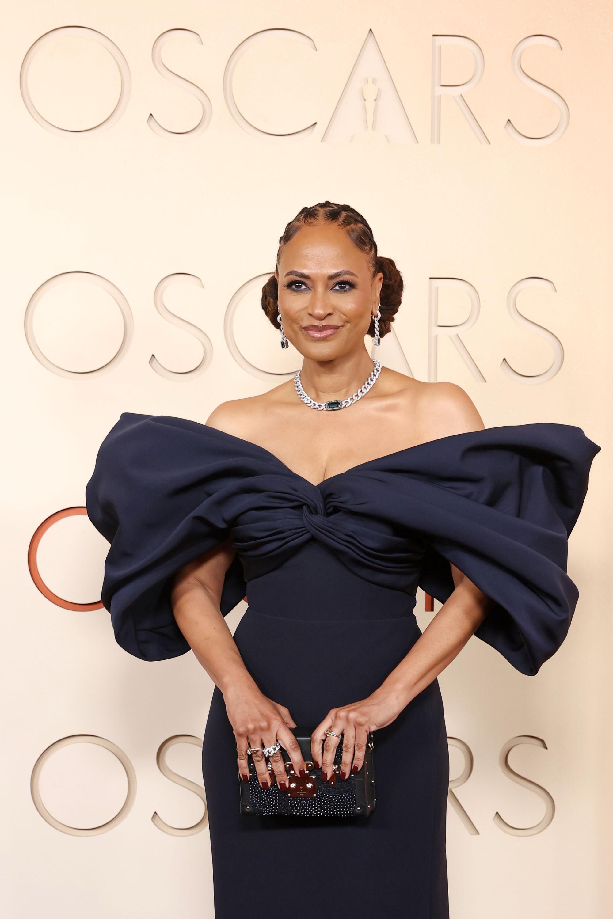 Ava DuVernay, wearing a black dress with exaggerated shoulders, poses on the red carpet. 