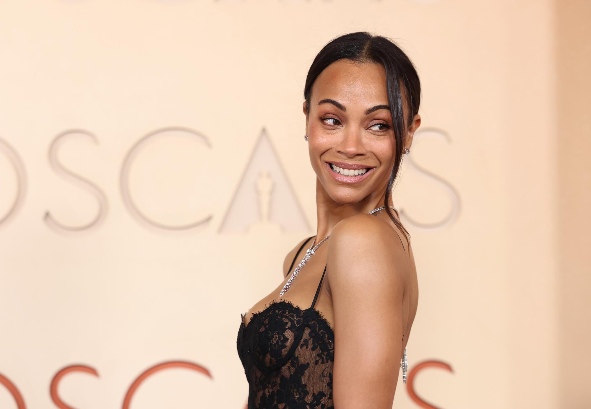 Zoe Saldaña, wearing a black lace dress, poses on the red carpet. 