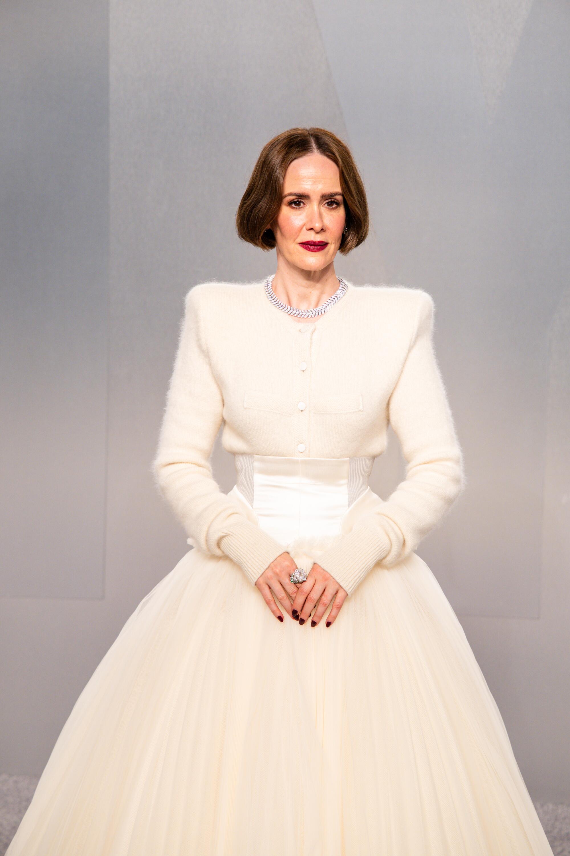 Sarah Paulson, wearing a white dress with a large tulle skirt, poses on the red carpet. 