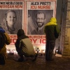 People wearing winter coats attend a candlelight vigil where Alex Pretti was killed in Minneapolis. Large posters of Alex Pretti and Renee Macklin Good are on a wall, and candles are on the ground in front of the posters.