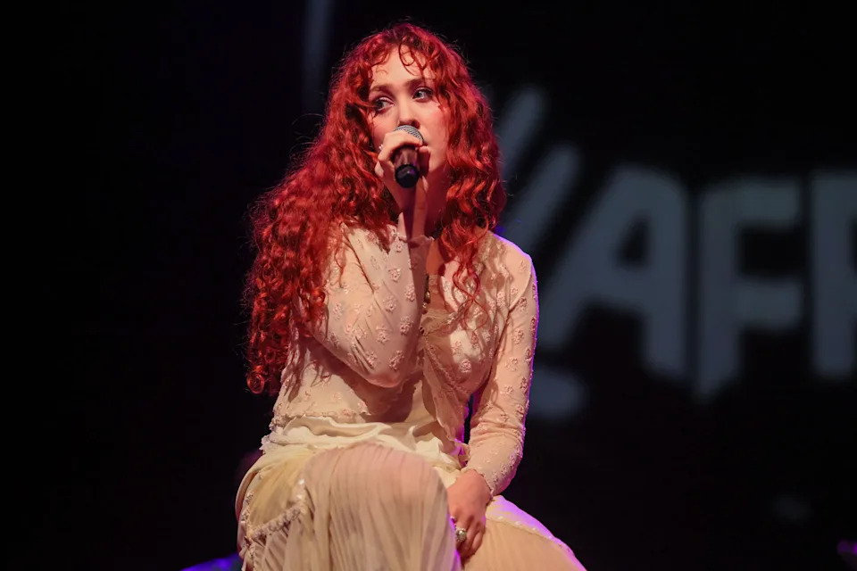 Performer with long, curly hair sings into a microphone on stage, wearing a flowing, sheer outfit. The background is dark with indistinct text