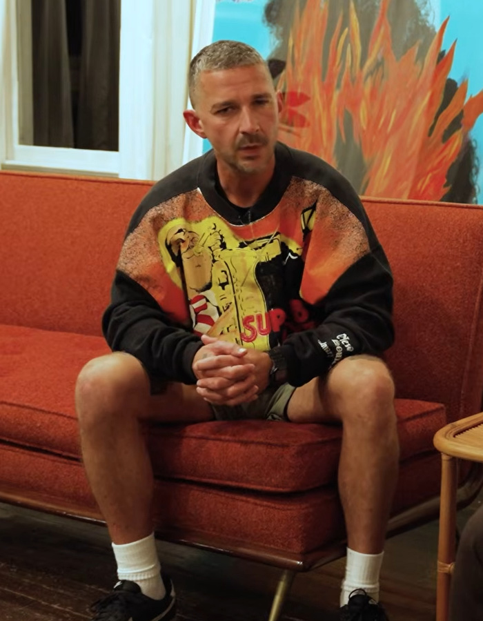 Shia LaBeouf sitting on a couch, wearing a colorful sweatshirt, discussing his Mardi Gras arrest and mental health struggles.