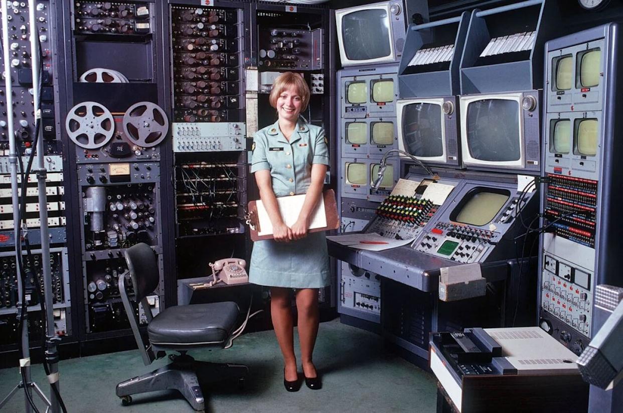 A woman in an Army uniform stands in front of a bays of televisions and audio devices -