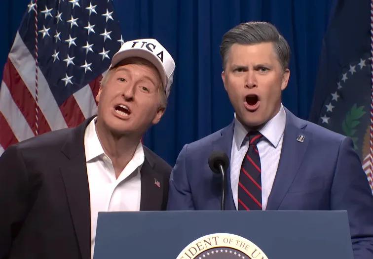 Two performers mimic a political scene on stage, standing at a podium with flags in the background. One wears a "USA" cap