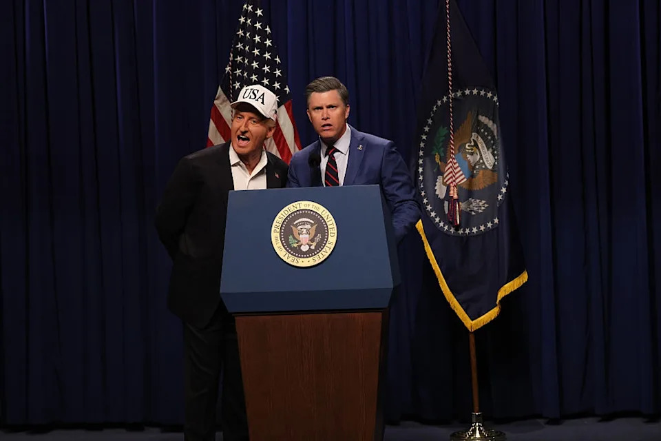 Two actors impersonating political figures stand at a podium labeled with a presidential seal, with USA and flags in the background