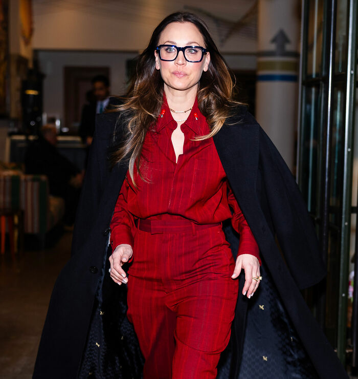 Kaley Cuoco wearing glasses and a red outfit with a black coat, walking confidently indoors calling out former co-star. Kaley Cuoco wearing glasses and a red outfit with a black coat, walking confidently indoors calling out former co-star.