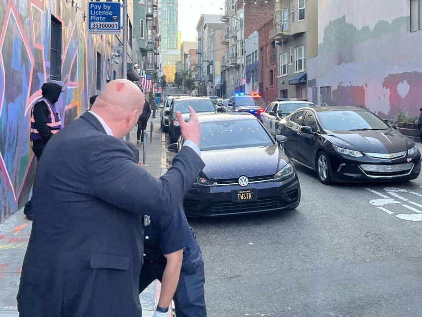 Police officers and a suited individual stand in an urban alleyway with police cars and flashing lights, while several bystanders observe the scene.
