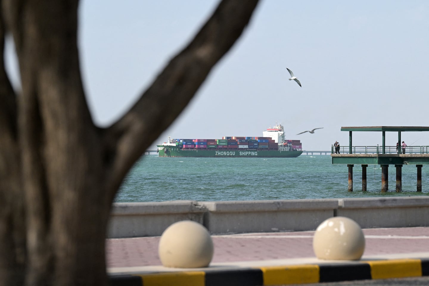 A Zhonggu Shipping container ship sails past people fishing on a pier on the Sheikh Jaber al-Ahmad al-Sabah Causeway on its way towards the port of Shuwaikh in Kuwait City on February 28, 2026. 