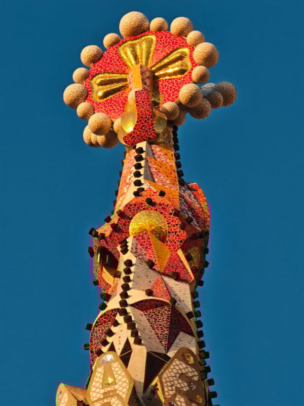 Colorful mosaic tower with a circular, flower-like top adorned with spherical shapes, set against a clear blue sky. The structure features red, yellow, and gold tiles arranged in intricate patterns.