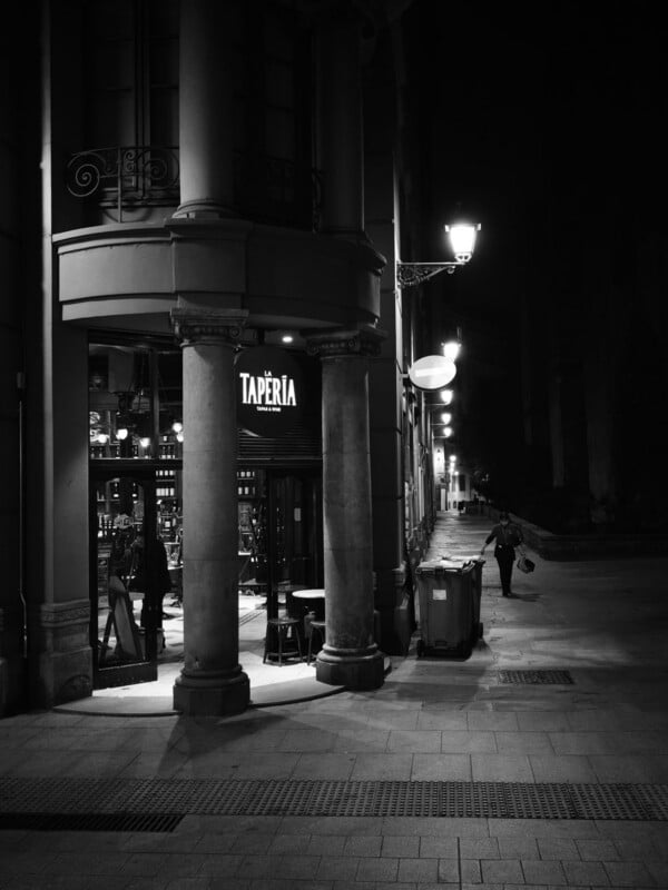 Black and white photo of a street at night, showing the entrance of a restaurant called "Tapería" with tables outside. Street lamps light the sidewalk, where a person walks past a trash bin further down.
