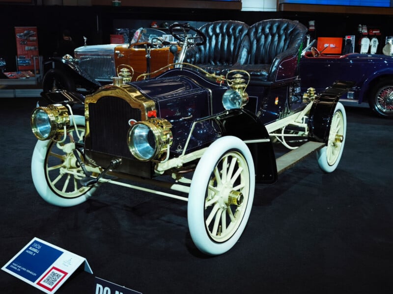 A vintage 1912 electric car with white wheels, brass headlights, and a dark body is displayed indoors on a black carpet, with an information plaque in front and other classic cars in the background.