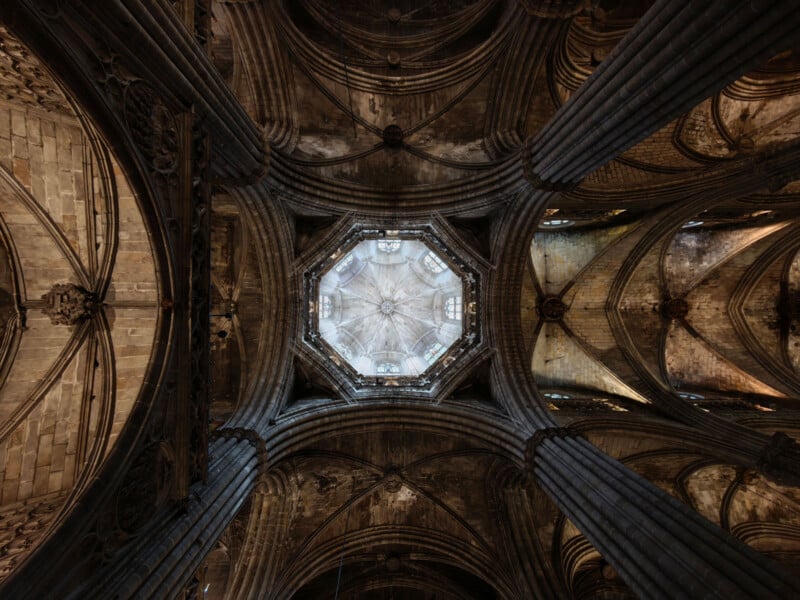 View looking up at an ornate, octagonal dome ceiling in a Gothic cathedral, surrounded by tall, ribbed stone columns and arches, with intricate architectural details and muted, earthy tones.