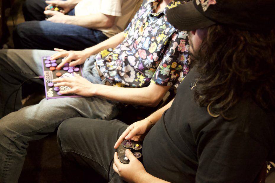Two people are seated side by side, one using a game controller and the other using a fight stick to play a video game.