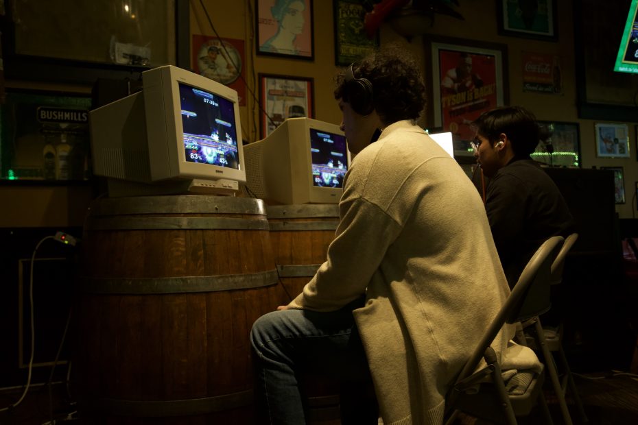 Two people sit on chairs facing old CRT monitors on barrels, playing video games in a dimly lit room decorated with posters.