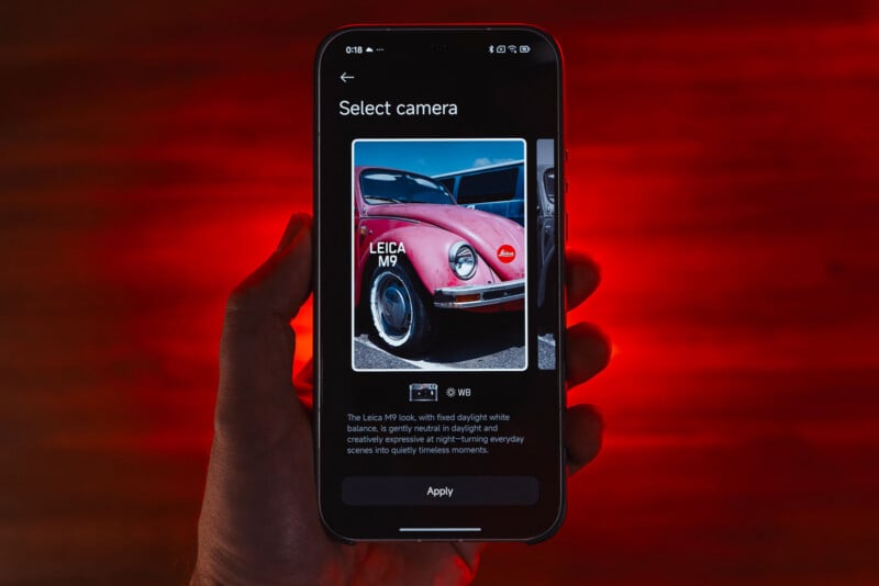 A hand holds a smartphone displaying a camera selection screen. The screen shows a close-up photo of a classic red car with the label “LEICA M9,” and an “Apply” button at the bottom. The background is dark with red lighting.
