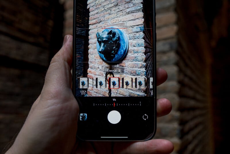 A hand holds a smartphone, taking a photo of a sculpture of a bull’s head mounted on a brick wall. The phone screen displays the camera app with various filter options visible below the image.