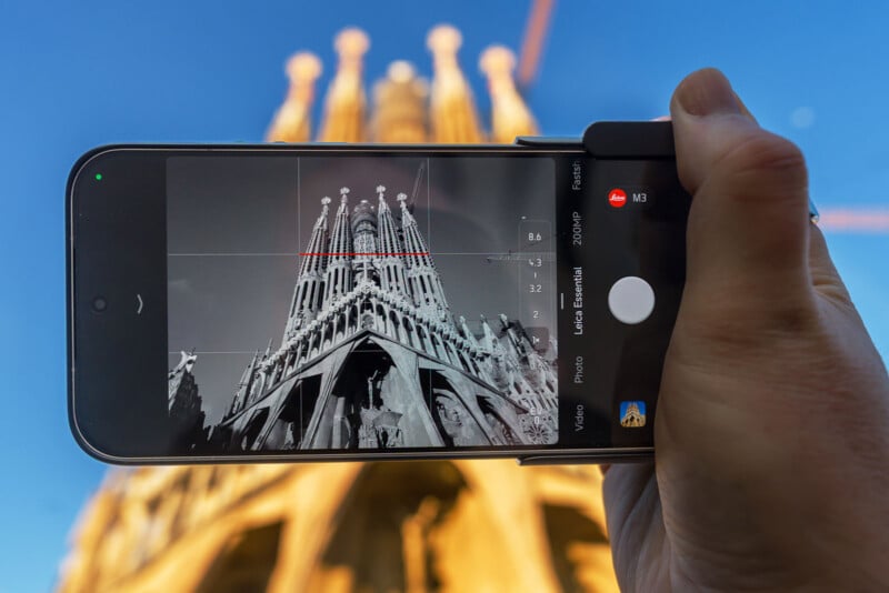 A hand holds a smartphone, taking a black-and-white photo of the Sagrada Familia’s ornate facade against a clear blue sky. The real basilica appears blurred in the background, while the phone screen shows it in sharp detail.