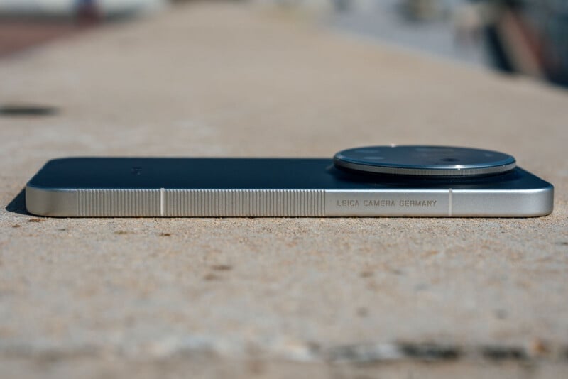 A close-up side view of a smartphone with a large protruding camera module, lying flat on a rough, beige surface. The words "LEICA CAMERA GERMANY" are engraved on the metallic edge.