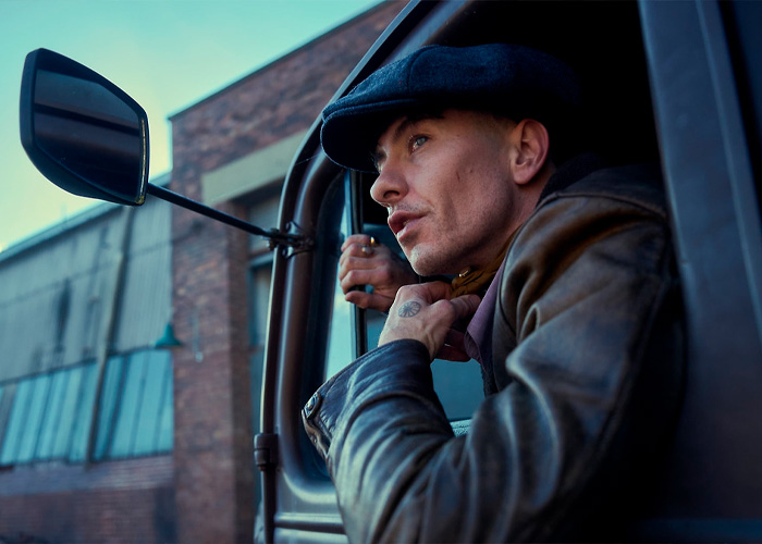 Barry Keoghan wearing a black cap and leather jacket, looking out from a vehicle against an urban background