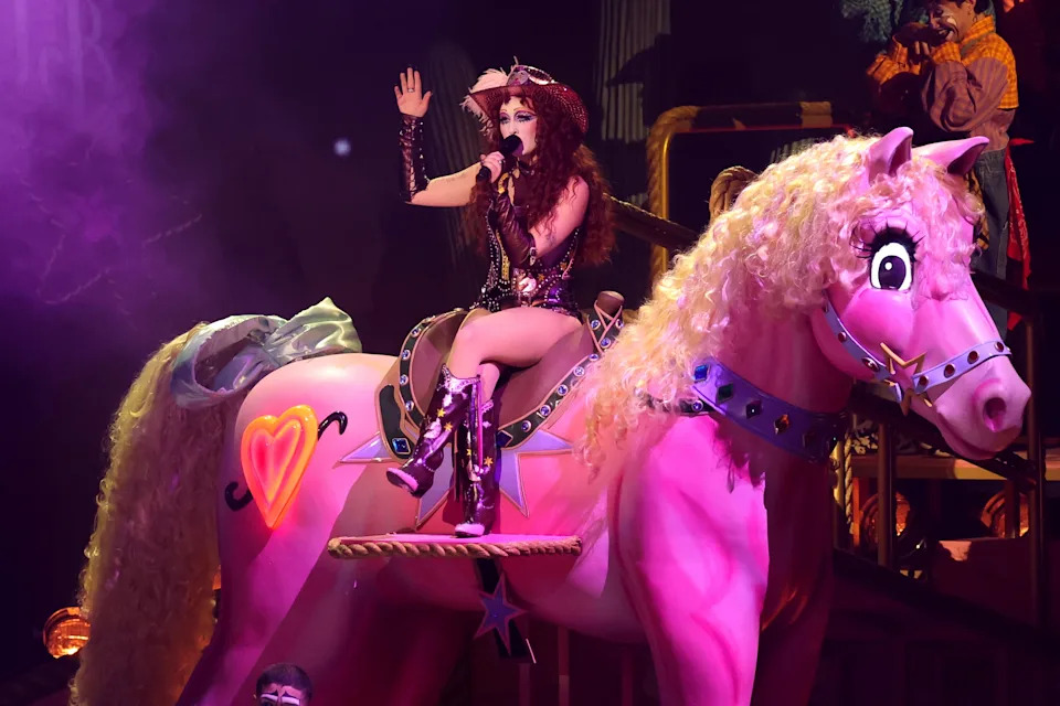 Singer performing on stage, dressed in theatrical, embellished attire while riding a large, decorative horse prop