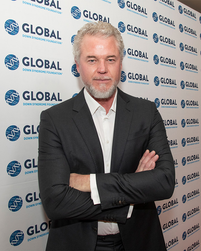 Man in black suit with gray hair and beard standing with arms crossed at a Global Down Syndrome Foundation event.