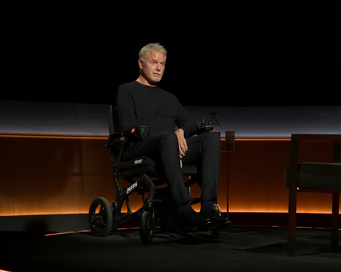 Man in black sitting in a wheelchair on a dark stage with dim orange lighting, relating to Eric Dane’s cause of passing.
