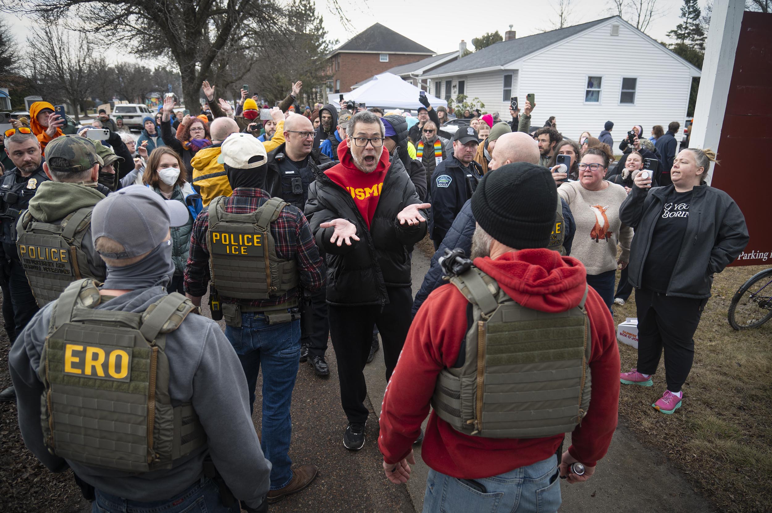A group of people confront police and ICE officers wearing tactical vests during a tense outdoor encounter in a residential neighborhood.