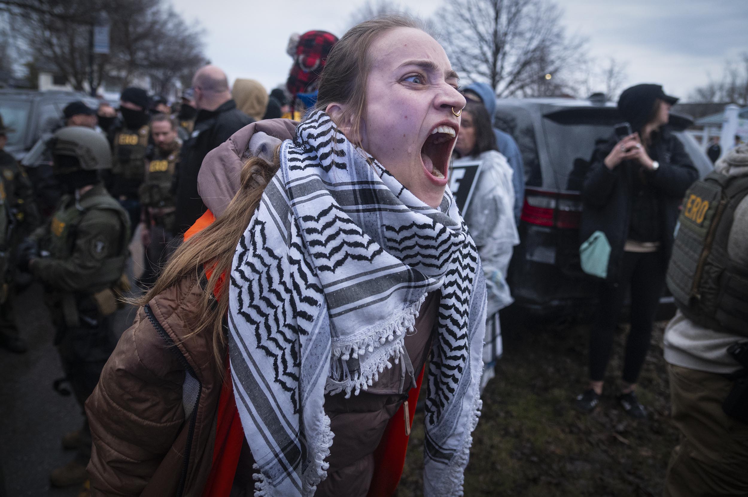 A person wearing a patterned scarf and orange vest shouts amid a crowd of people, some in tactical gear, during an outdoor protest or demonstration.