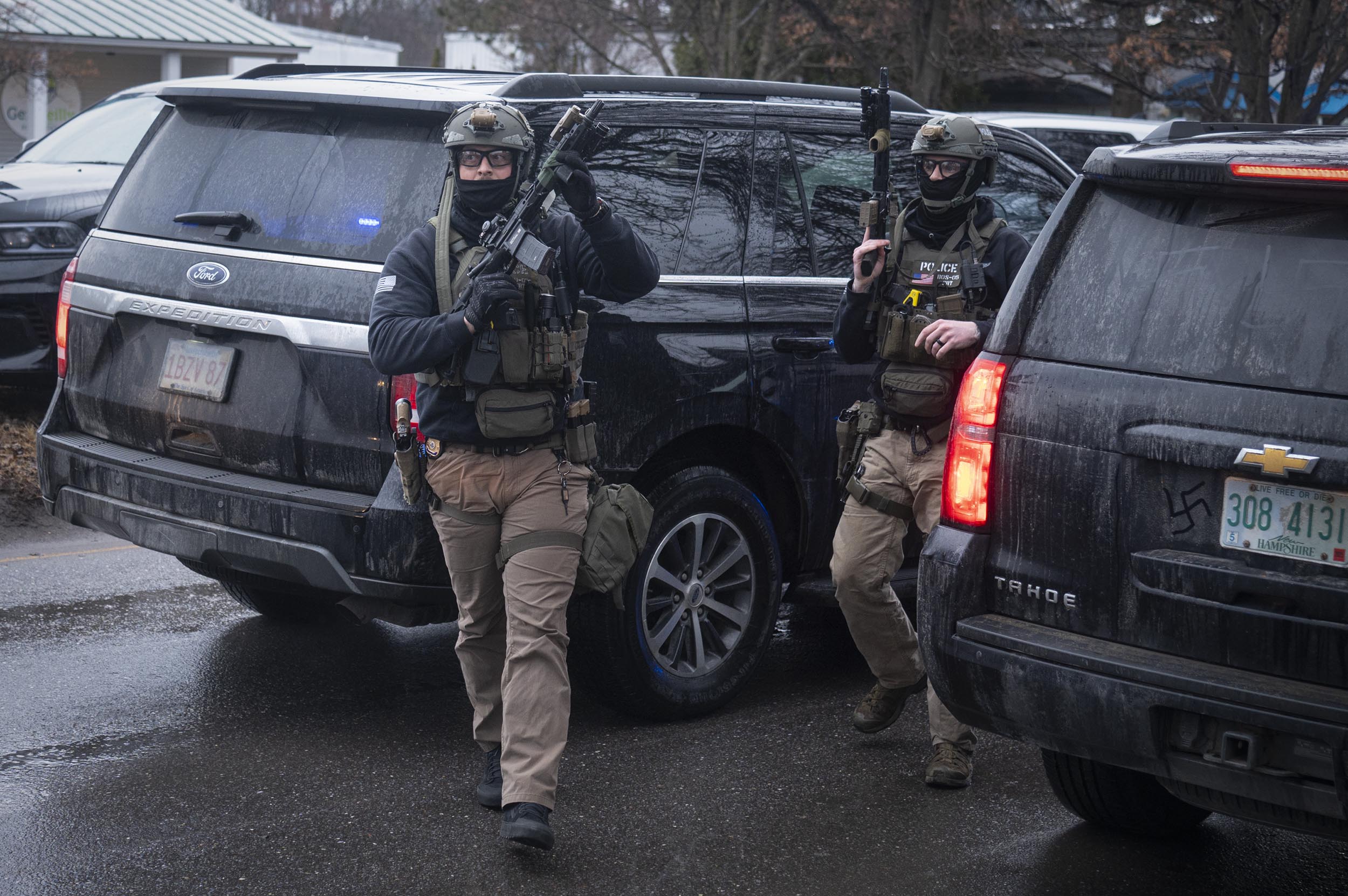 Two armed law enforcement officers in tactical gear walk between SUVs on a wet street, appearing to respond to an active situation.