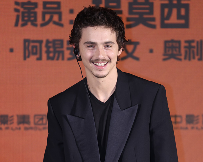 Actor at an event smiling, dressed in a black suit jacket, with an orange background featuring text in another language.