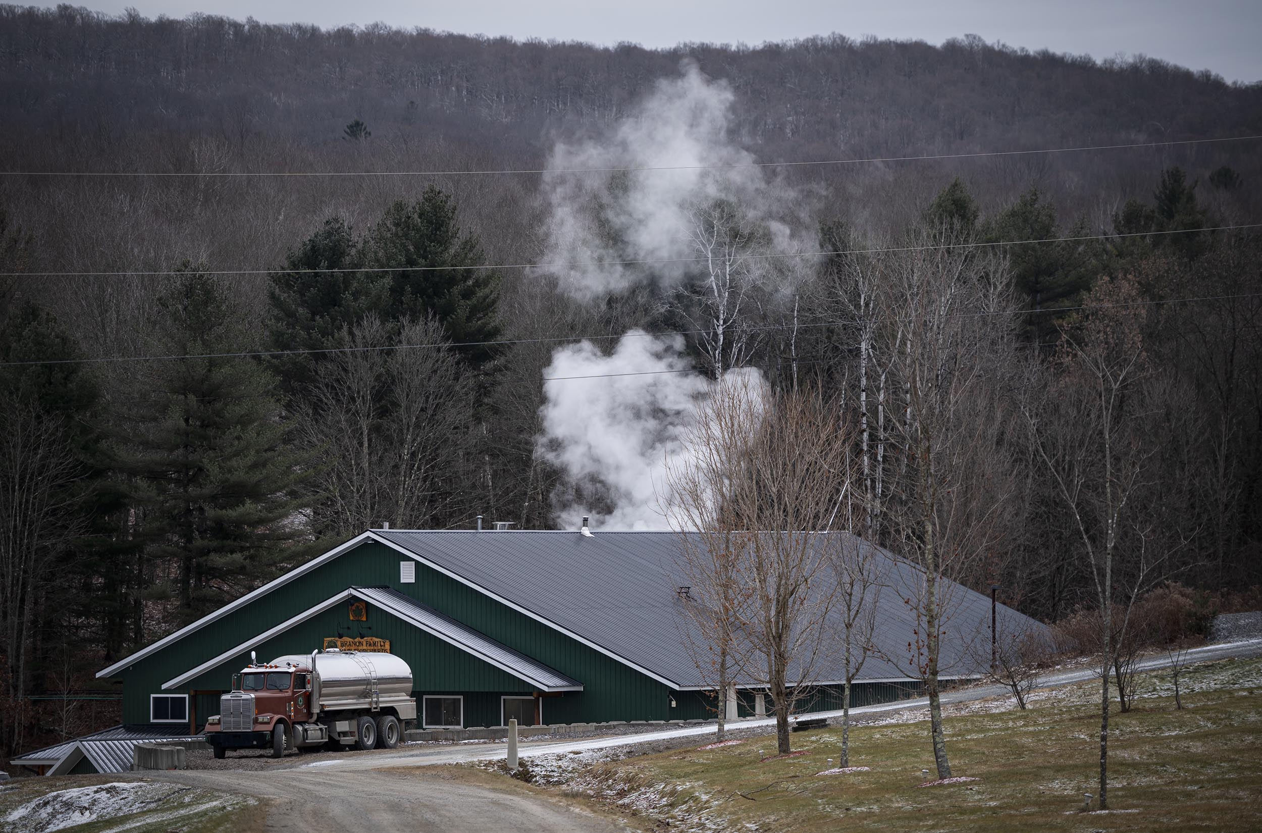 A green building with smoke rising, surrounded by bare trees and hills. A tanker truck is parked on a dirt road nearby.