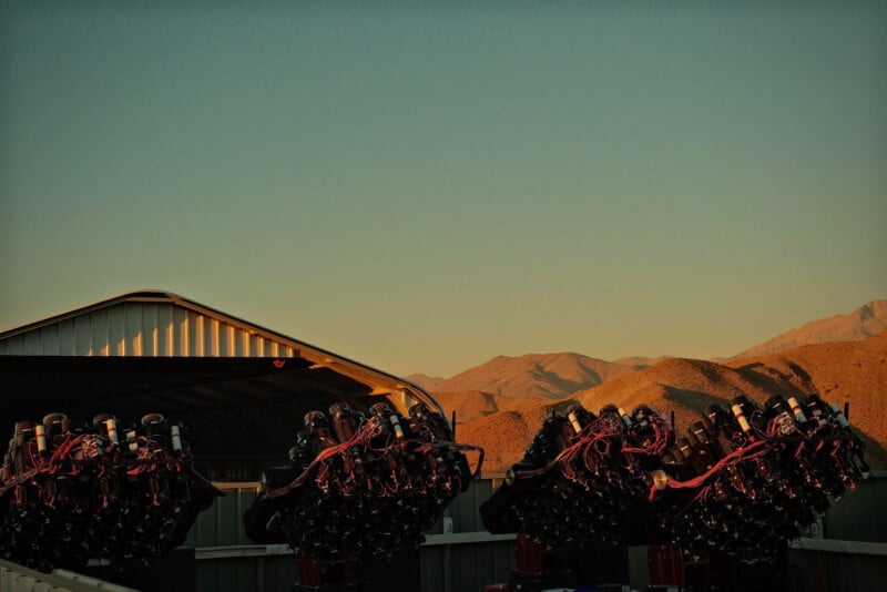 A row of high-tech telescope arrays stands outside a metal shed at sunset, with mountains and dry terrain in the background under a clear sky.