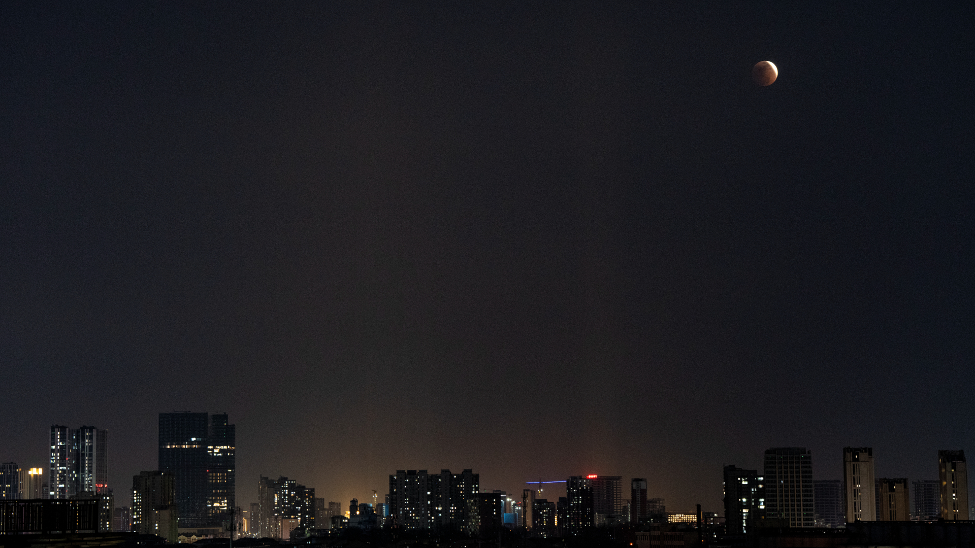 An almost fully eclipsed moon hangs low over a city skyline at night, with a thin crescent of its upper right edge illuminated by sunlight.