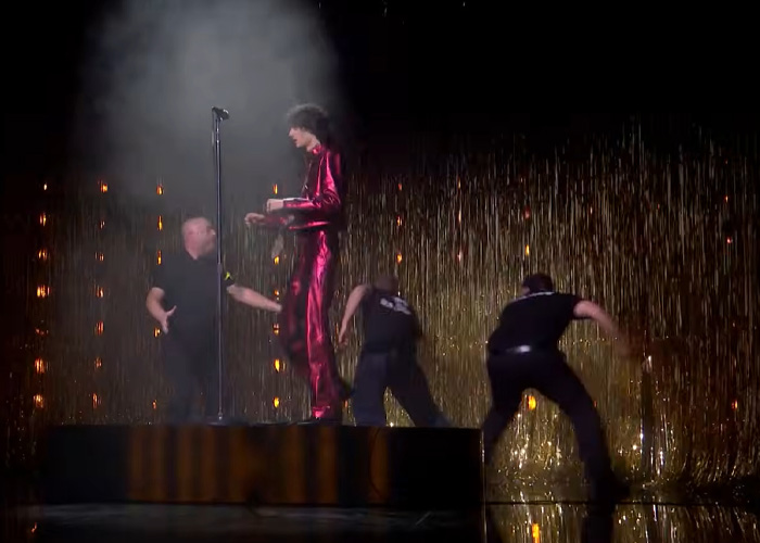 Singer in red outfit being pushed off stage during chaotic Brit Awards performance with security and gold curtain backdrop. Singer in red outfit being pushed off stage during chaotic Brit Awards performance with security and gold curtain backdrop.