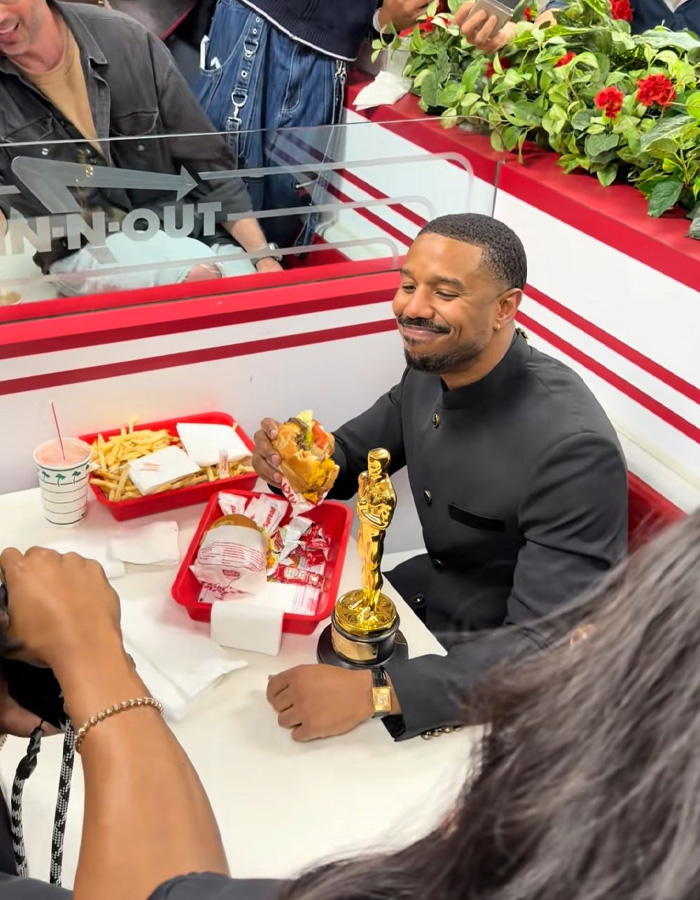 Michael B. Jordan smiling and holding an Oscar statue while eating a burger at In-N-Out fast food restaurant.