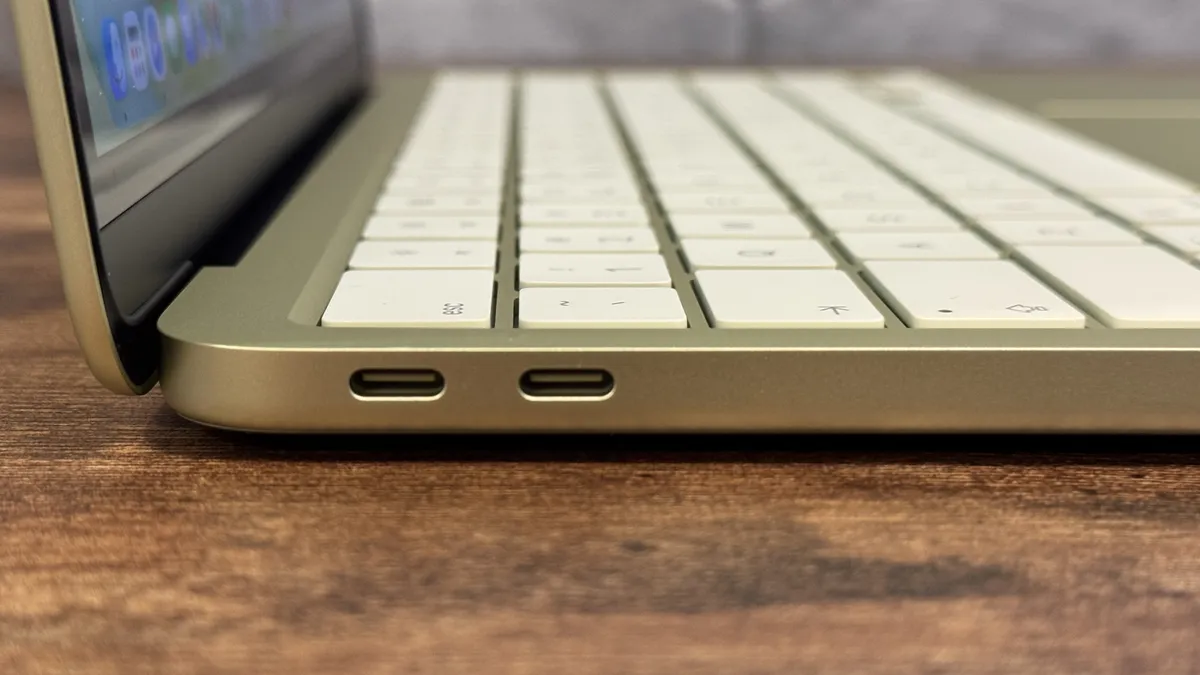 Slim laptop on a wooden surface, side view showing two USB-C ports. The keyboard is visible, with a glimpse of the screen displaying blurred icons.